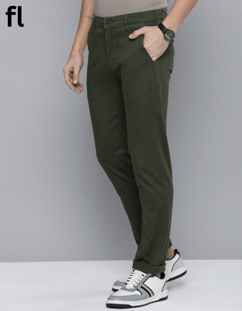 A man standing against a white background, wearing olive green cotton pants and a grey shirt, with a jacket in his hand and white sneakers on his feet.